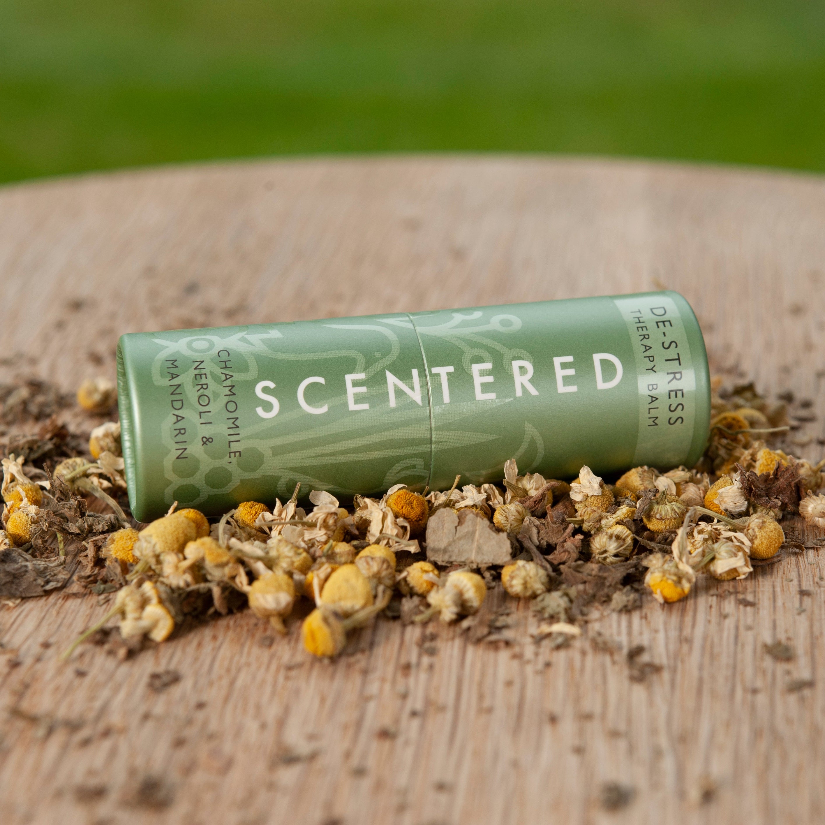 This is a picture of a Scentered De-stress Aromatherapy Balm, full size 5g, lying on its side on a table surrounded by the ingredients that are included in the balm, especially chamomile