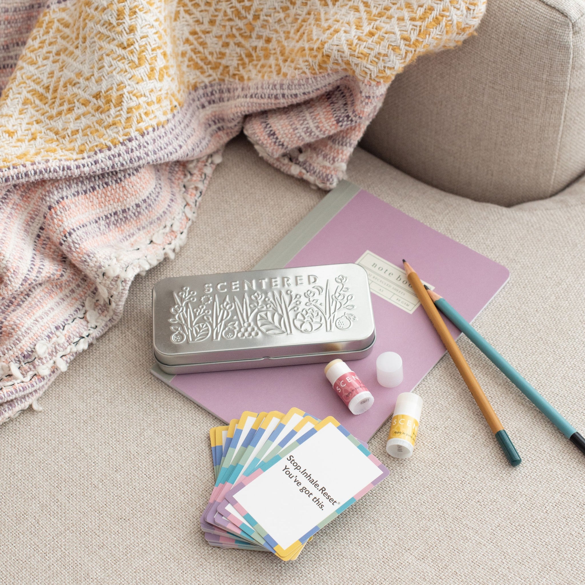 A tin box, a pack of cards, two mini balms , a book and pencils are resting on the sofa.