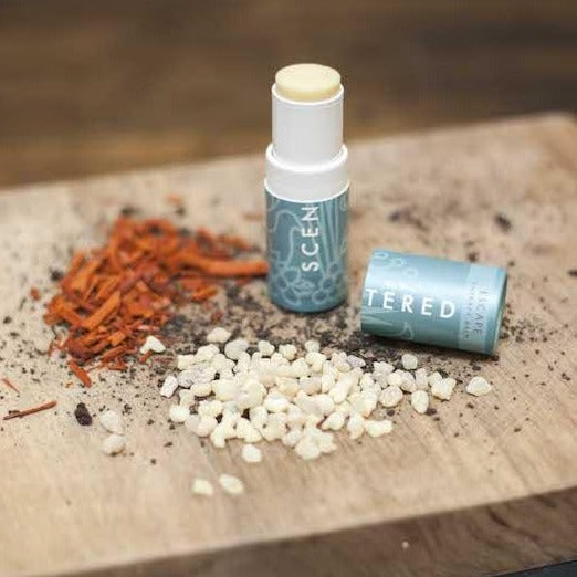 Scentered Escape Aromatherapy Balm, open, on wood surrounded by the fresh ingredients that are included in the balm, like oud and frankincense