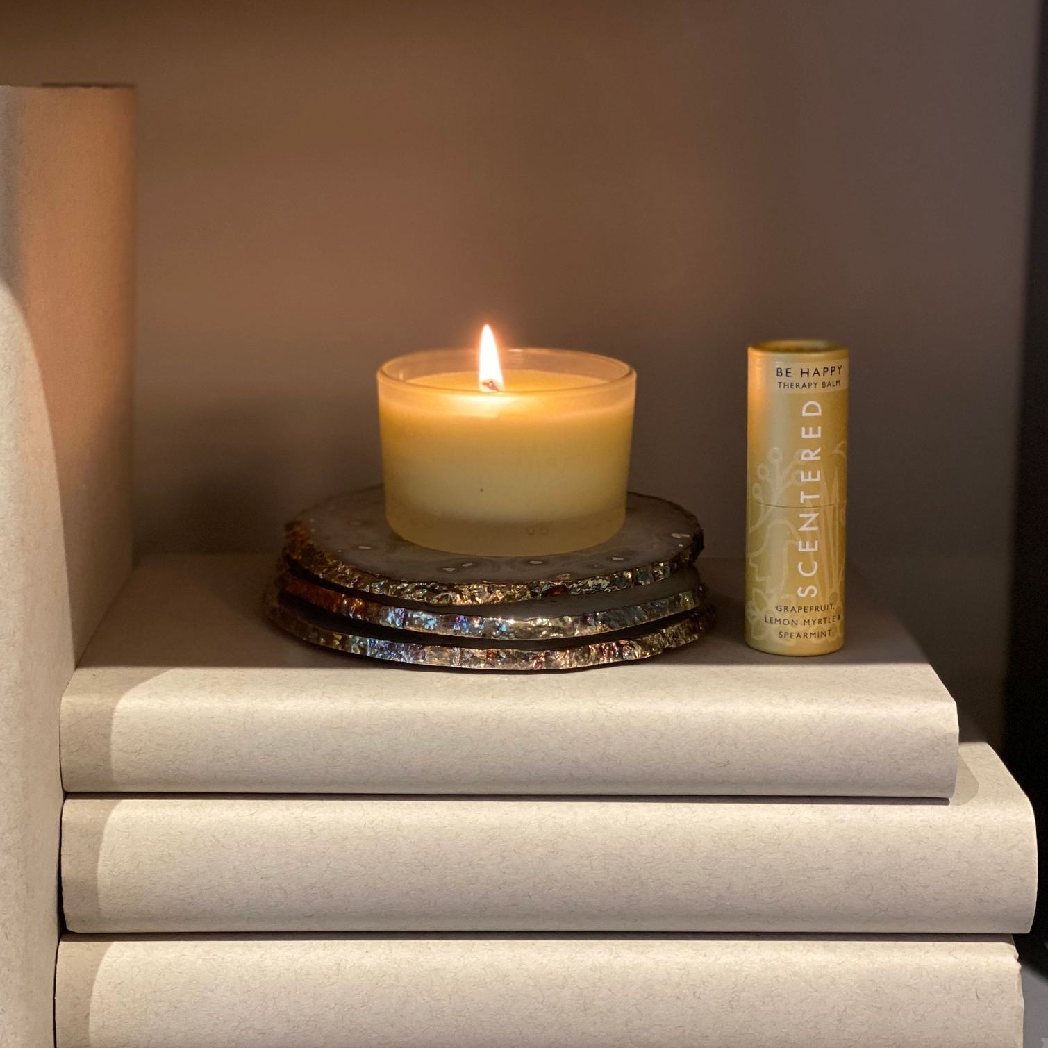 A lit Scentered Happy Aromatherapy Travel candle on a shelf next to the Scentered Happy Aromatherapy Balm, uplifting aromatherapy balm stick by Scentered, formulated to enhance mood and promote positivity throughout the day.