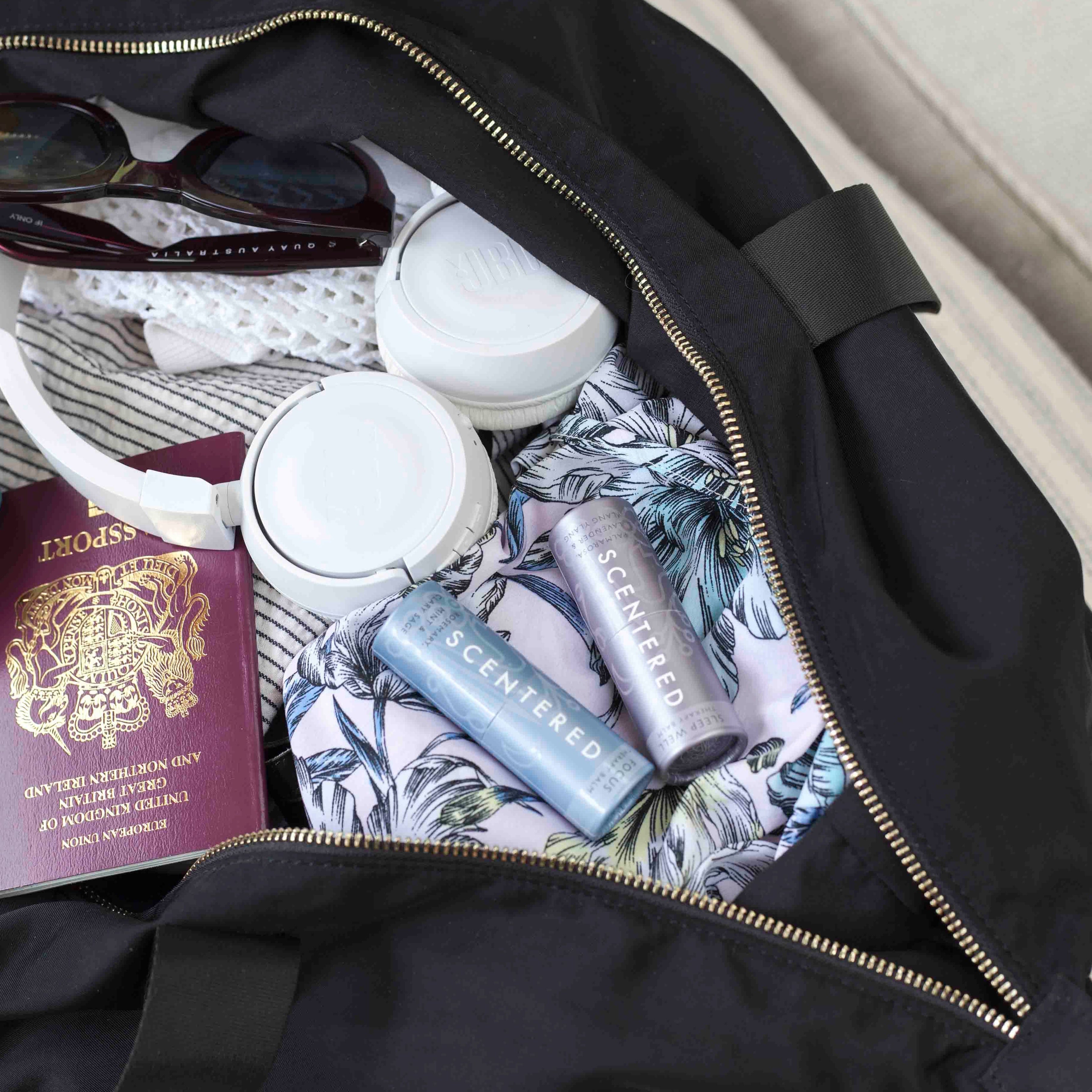 Scentered Sleep Well and Focus Aromatherapy Balms in black travel bag with passport, headphones, and sunglasses. Scentered Aromatherapy Balms - Sleep Well and Focus Travel Companions