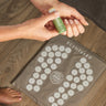 Acupressure Foot Mat on a wooden floor women's foot pictured in bottom left along with the woman applying the Scentered De Stress Balm to her wrist 
