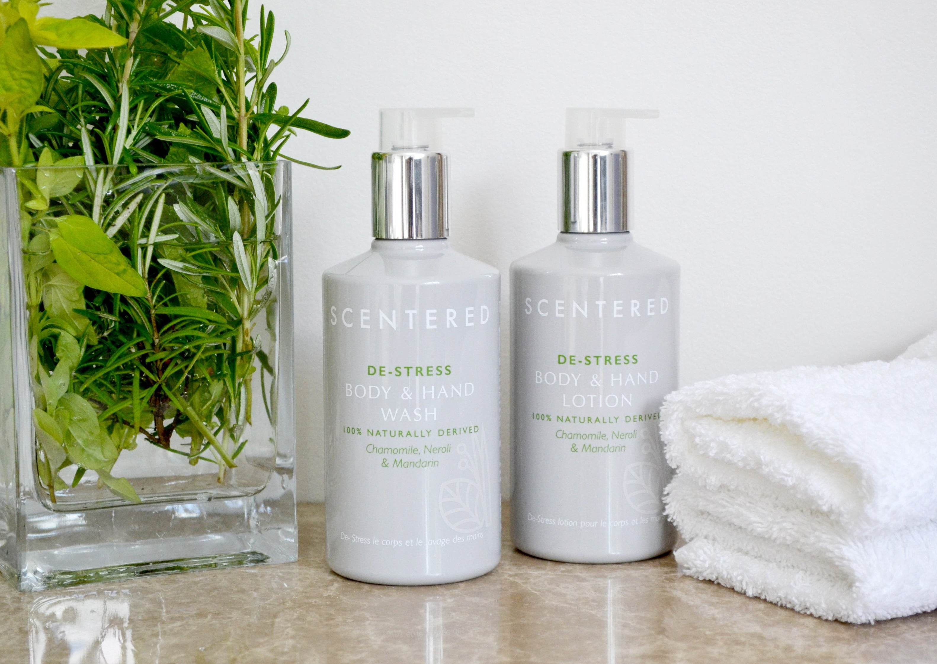 De-stress Wash and Lotion bottles on marble counter next to white towels and fresh herbs. Body Wash with Aloe and Lotion with Jojoba for Moisturized Skin.