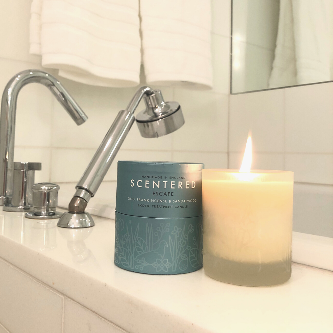 A lit Escape home candle next to its packaging by the bath tub