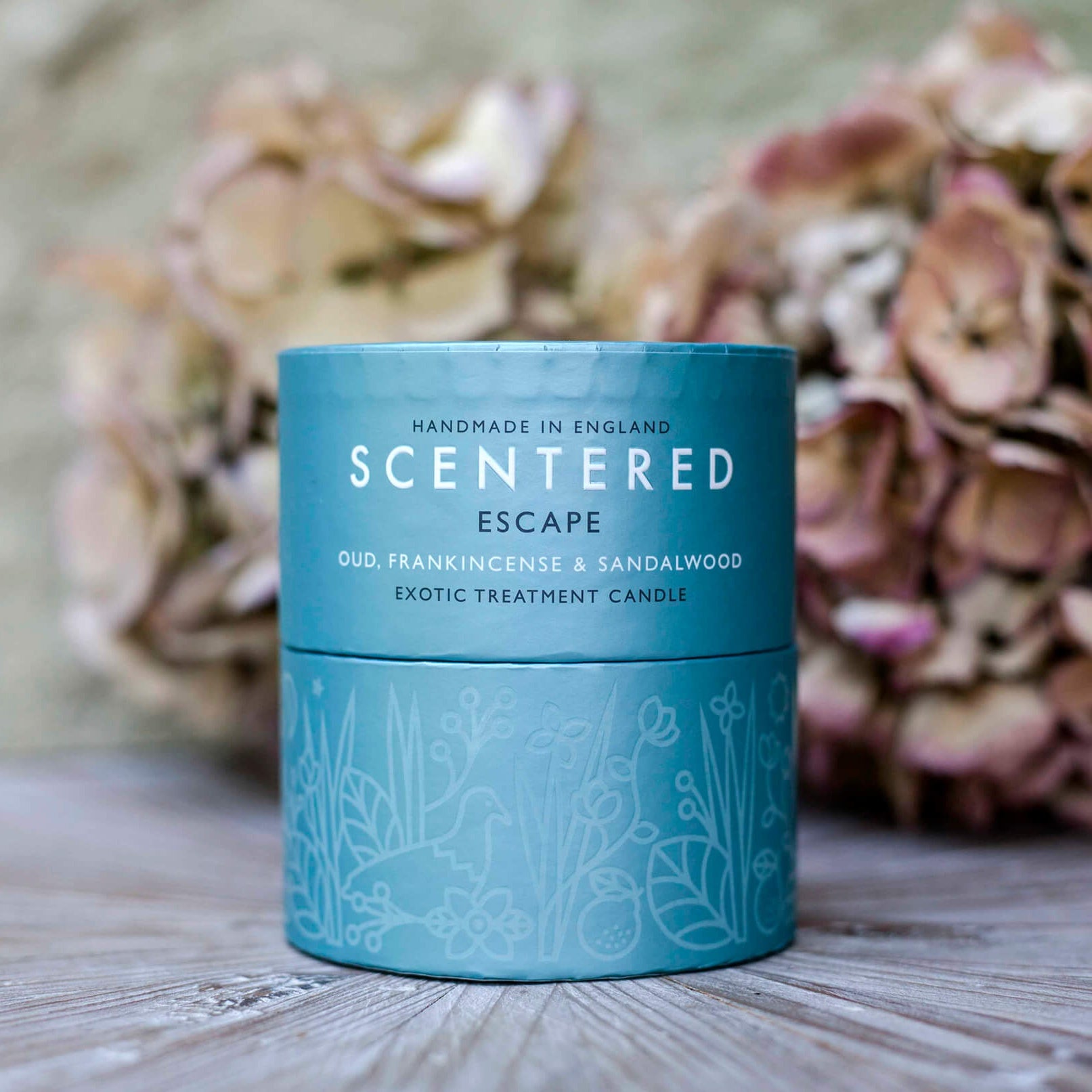 Escape home candle in its packaging with dried flowers out of focus in the background. Aromatherapy Candle for Wellness.